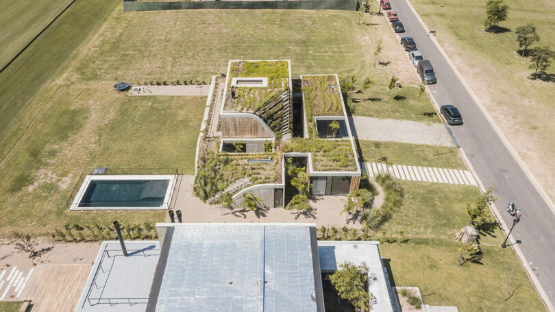 Aerial view of a modern, eco-friendly house with a green roof, a garden, and a rectangular swimming pool, situated near a paved road with parked cars and surrounded by grassy areas.