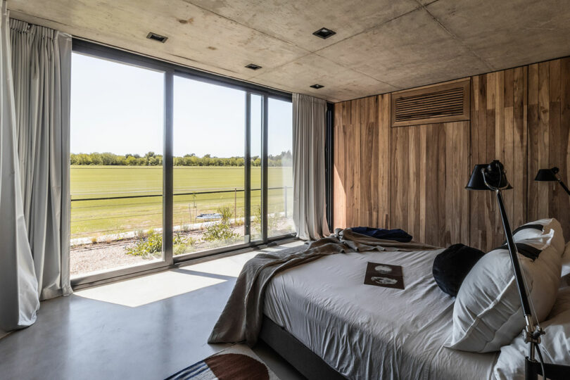 Modern bedroom with large windows, wooden walls, and a view of a green field.