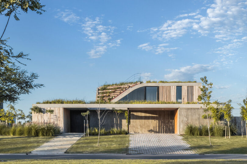 Modern concrete house with large windows and a green roof, surrounded by grass and trees, under a blue sky with scattered clouds.