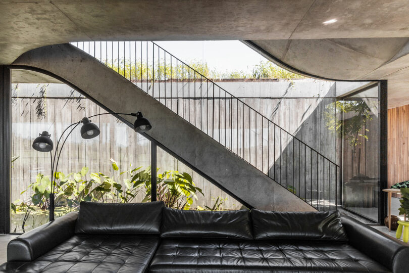 Modern living room with a black leather sofa, concrete walls, large windows, and a sleek metal staircase, featuring greenery visible through the glass.