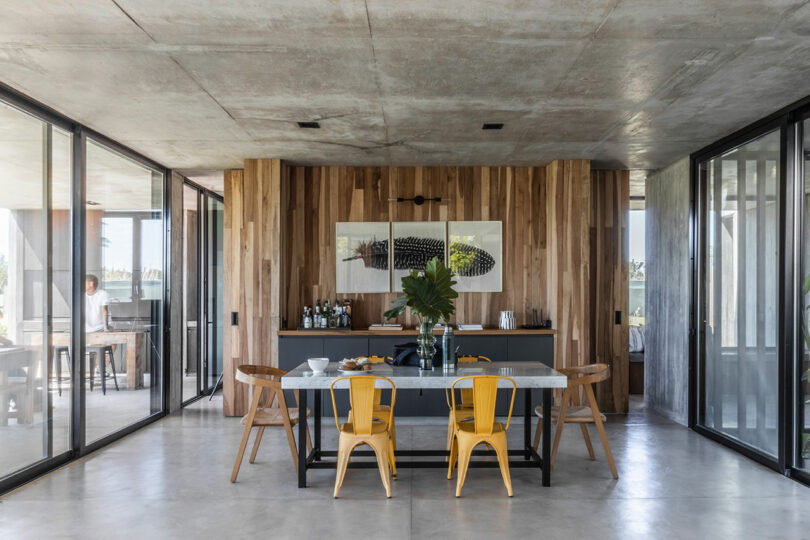 Modern dining room with a marble table, yellow chairs, and a wooden accent wall. Large glass doors and windows provide natural light. Two artworks hang on the wall.