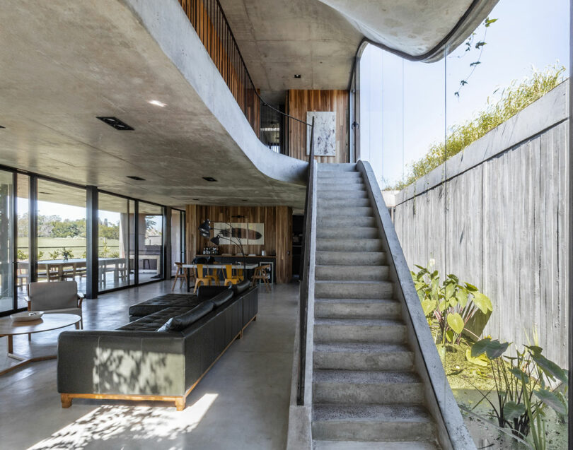 Modern interior with concrete staircase, large windows, a green sofa, wooden accents, and open space design. Natural light illuminates the area.