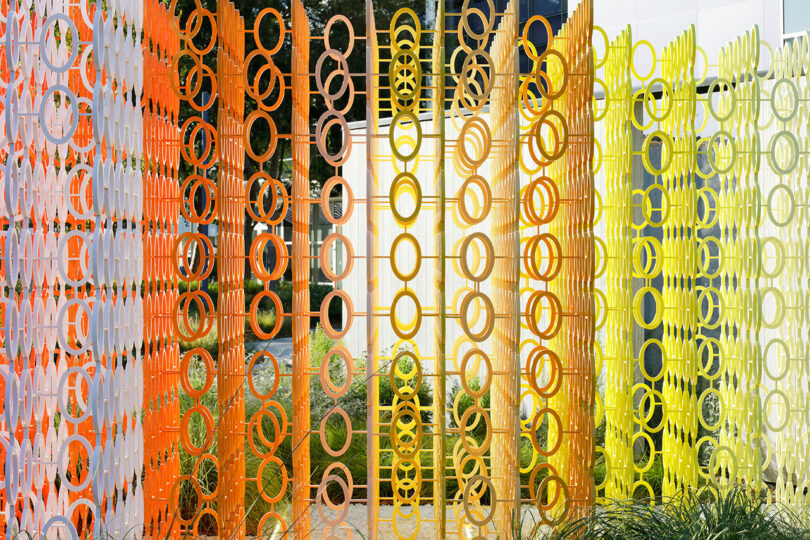 Colorful metal sculpture of interlocking circles in orange, yellow, and green shades forms a fence-like structure in front of a building, reminiscent of the vibrant style seen in Emmanuelle Moureaux's 100 Colors series.