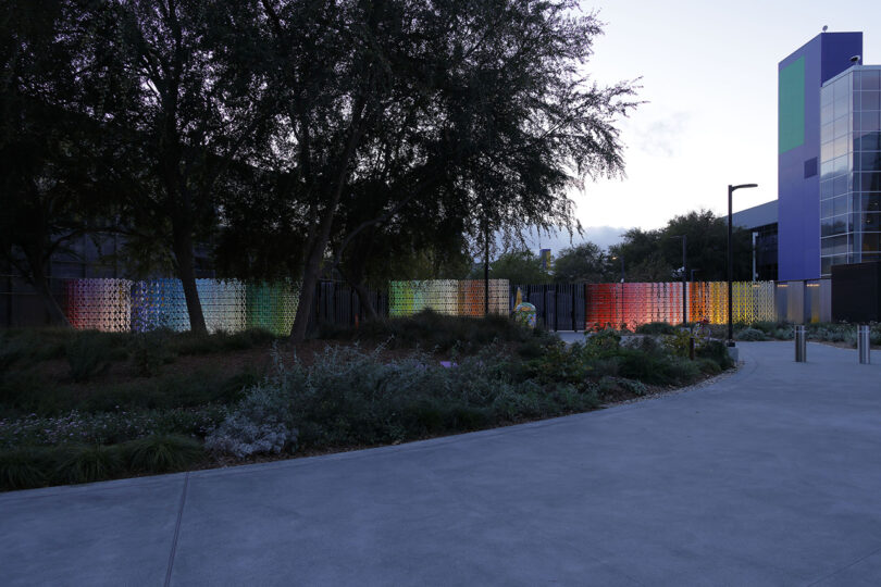 A row of colorful, translucent panels is displayed along a pathway surrounded by trees and plants, with a modern building in the background.