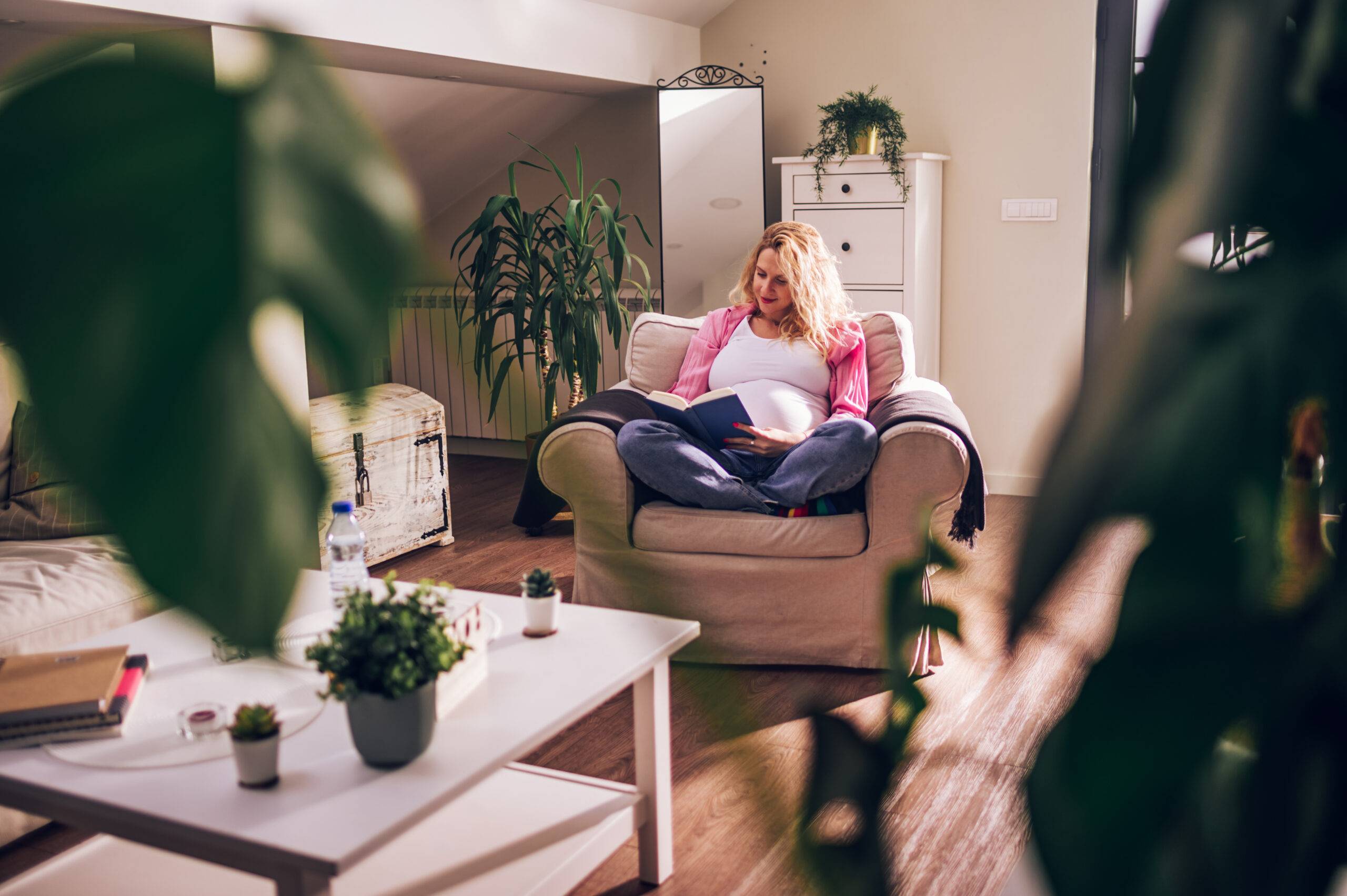 Young blonde pregnant woman reading a book for baby in belly. Future mother feeling happy while sitting on a chair in living room at home. Reading information about baby development.