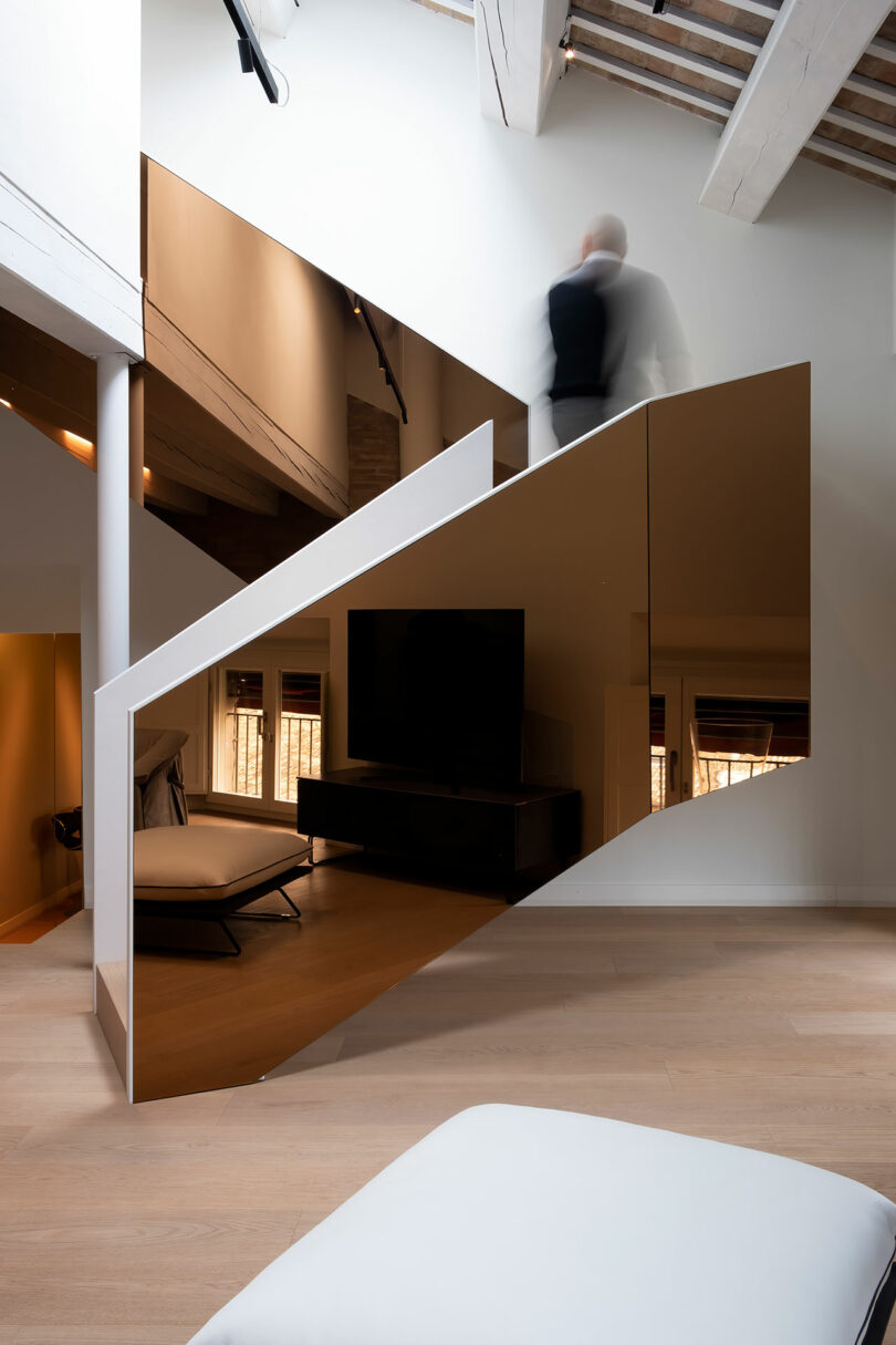 A modern interior with a mirrored staircase. A person walking up the stairs is blurred in motion. Minimalistic design features white walls, wooden floors, and a large TV.