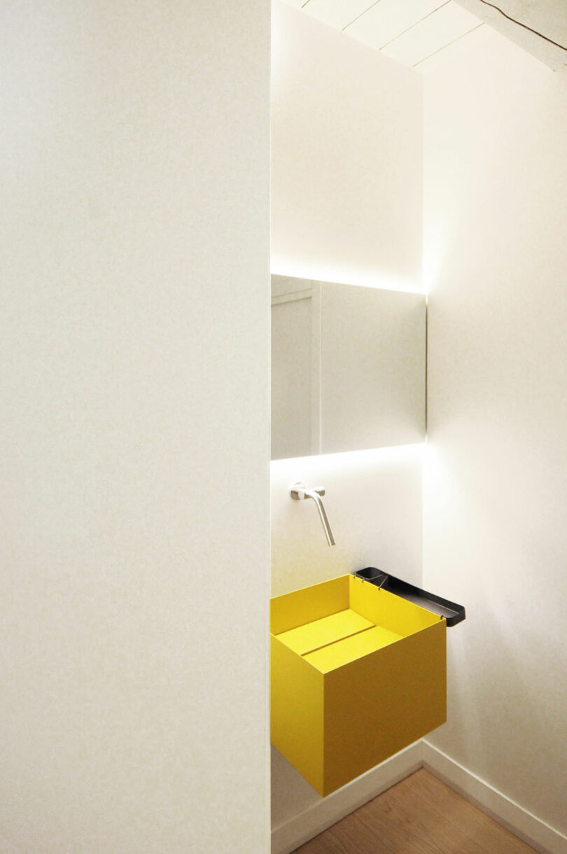 A minimalist bathroom features a rectangular yellow sink with a sleek wall-mounted faucet, a large mirror, and concealed lighting on a white wall.