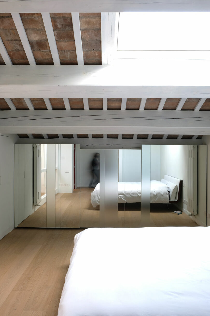 Bedroom with slanted wooden beams and a skylight, mirrored closet doors reflect a single bed with white bedding on wooden flooring, and a blurred figure is visible in the reflection.