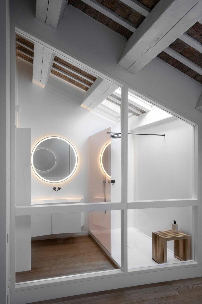 Modern bathroom with two circular, backlit mirrors on white walls, a large glass-enclosed shower, and a wooden stool on the floor. Exposed beams and a slanted ceiling add character.