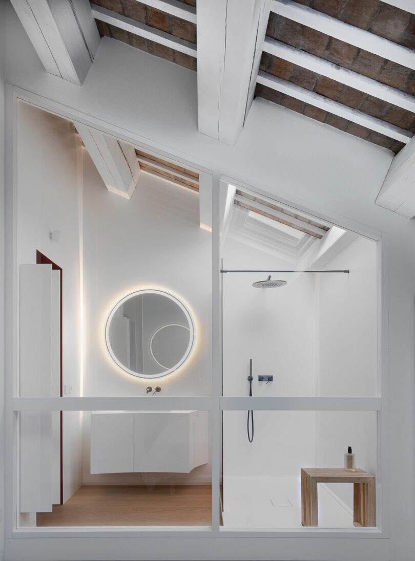 Modern bathroom with a round backlit mirror, a glass shower enclosure with a rainfall showerhead, and exposed beam ceiling. White walls and wooden accents are present.