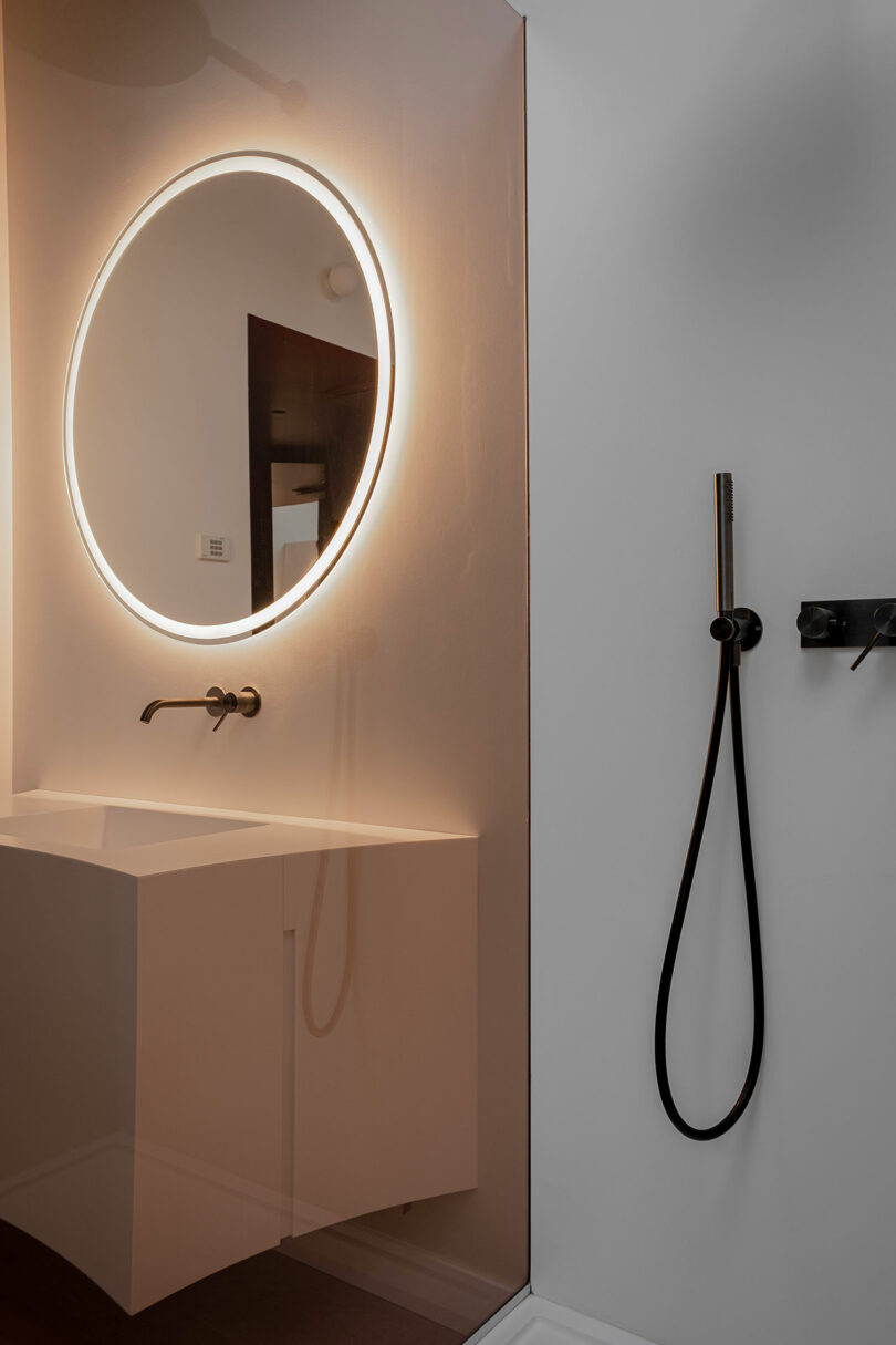 Minimalist bathroom with a backlit circular mirror above a white rectangular sink. A modern black shower fixture is on the adjacent wall.