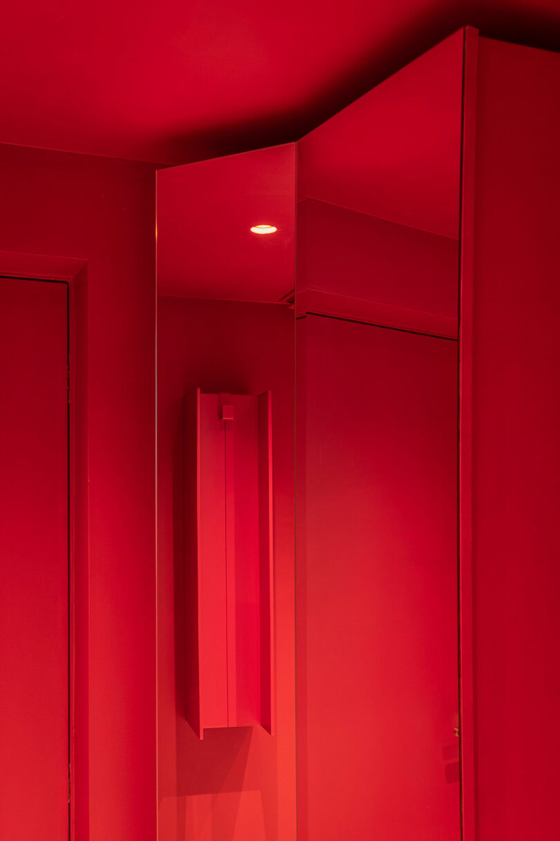 A room with red walls, a closed door, and a narrow rectangular wall fixture. A ceiling light illuminates the scene.