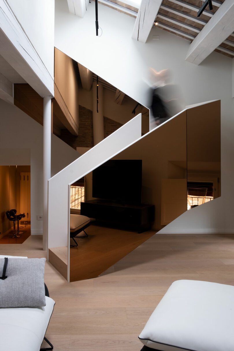 Triangular mirrored staircase in a modern, minimalistic room with wooden floors and exposed beams. A blurred figure is walking up the stairs.