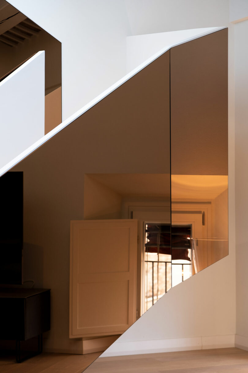 Modern interior with a staircase featuring a glass and metal railing, natural wood flooring, and a small window partially obscured by the structure.