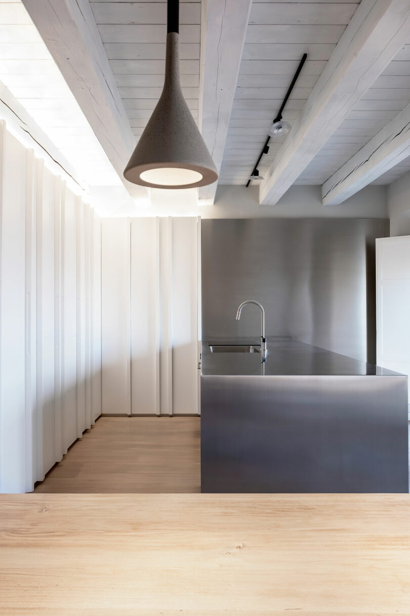 Modern kitchen with a stainless steel island, wooden floor, pendant light, and exposed beams. Minimalist design with white walls and ceiling.