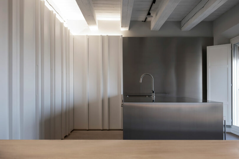 Minimalist kitchen with a stainless steel island and faucet, white panel cabinets, light wood countertop, and exposed wooden ceiling beams.