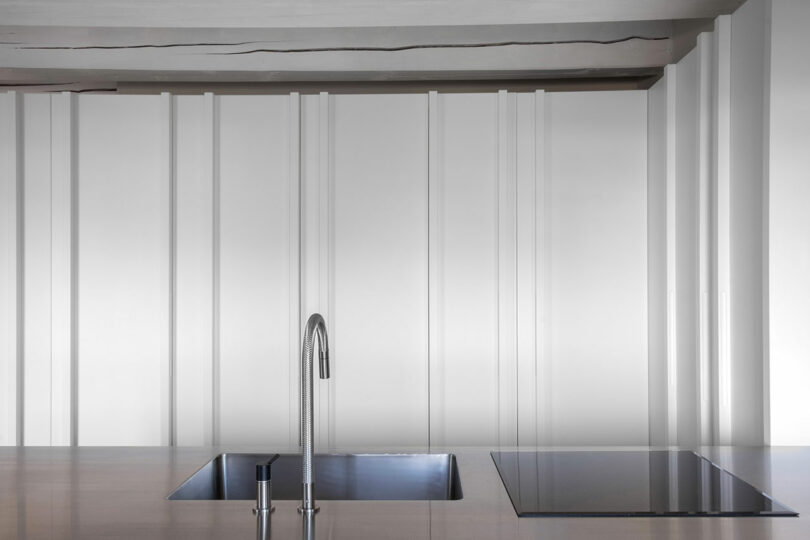 Minimalist kitchen with a sleek metal countertop featuring a modern faucet, a built-in sink, and an induction cooktop, set against a backdrop of vertical panel wall design.