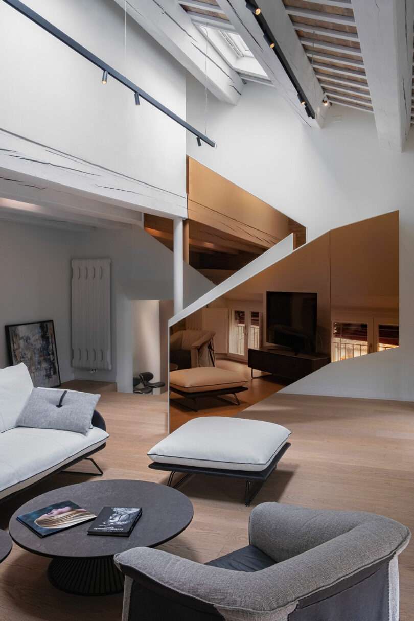 Modern living room with a minimalist design. Features include a large, angled mirrored wall, two contemporary sofas, a round coffee table, and exposed beams. Natural light illuminates the space.