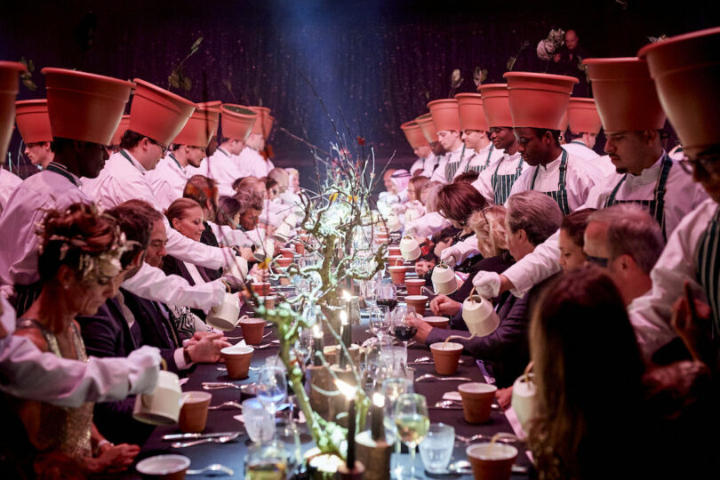 Charles Kaisin's Surrealist Dinner in 2019 - Metamorphosis in Mirano, Bruxelles. The image features guests sitting along both sides of a dining table, which is extravagantly decorated, with servers wearing terra cotta planters on their heads like a hat, pouring tea into terra cotta planter cups for the guests.