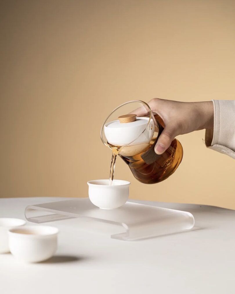 Teatsy Vortex Kit in Classic Crystal - features a hand pouring tea from a 360° rotational strainer into a white tea cup on a clear decorative stand 