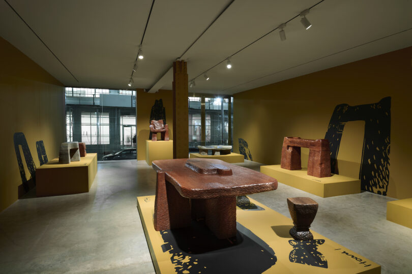 Museum exhibit with wooden and stone sculptures on platforms in a dimly lit room. Modern design with large windows and track lighting