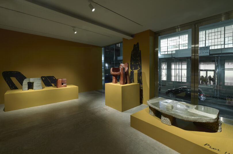 Exhibition room featuring abstract sculptural furniture on raised platforms against a yellow wall, illuminated by overhead lights