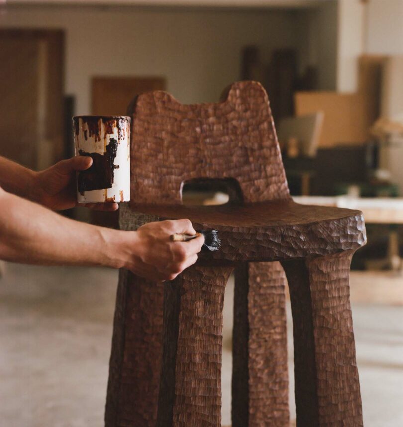 Person applying varnish to a textured wooden chair with a brush inside a workshop