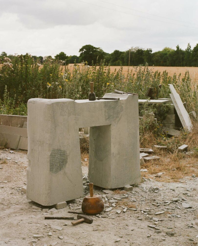 Stone block with a rectangular cutout stands on rocky ground, surrounded by tools and debris. Field and trees are visible in the background