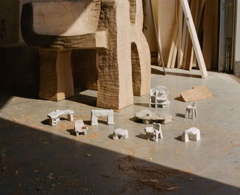 A collection of small, handcrafted clay furniture models are arranged on a sunlit concrete floor near a large unfinished wooden sculpture. Shavings are scattered around