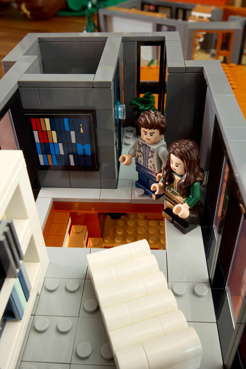 Two LEGO figures stand in a modern, open-plan LEGO room with abstract wall art and a white couch.