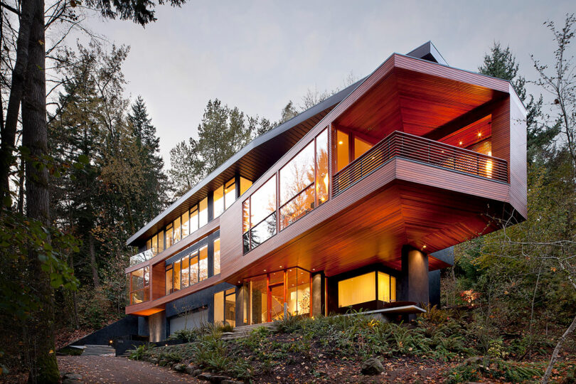 Modern angular house with large windows and wooden accents, set in a forested area. The house is elevated on pillars and surrounded by trees and greenery.