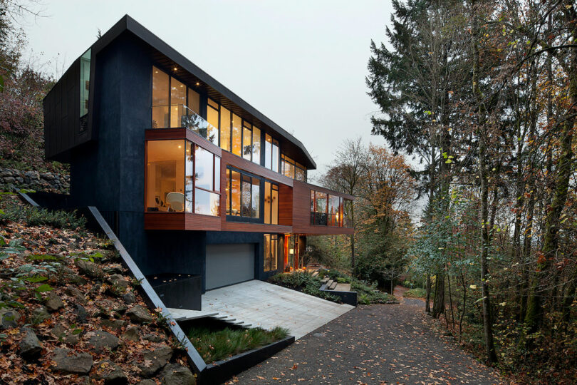 A modern, two-story house with large glass windows is set on a sloped, wooded lot with a concrete driveway.