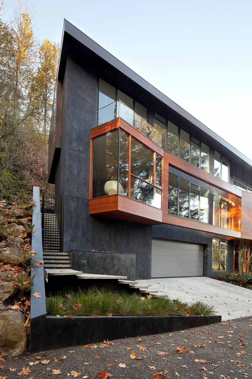 Modern two-story house with large windows, dark exterior, and wooden accents. Stairs lead up to the entrance beside a garage. Surrounded by trees and fallen leaves.