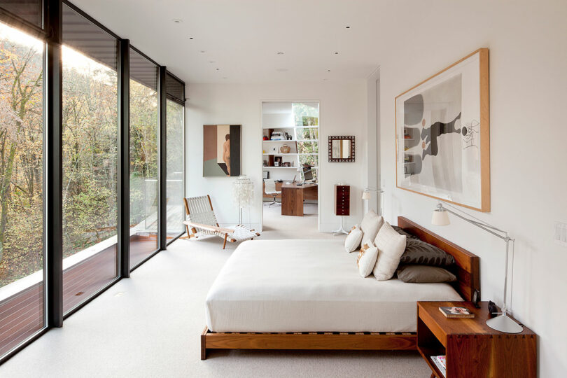 Modern bedroom with large glass windows, a wooden bed, and a view of trees. An adjacent open area features a chair and wall art.