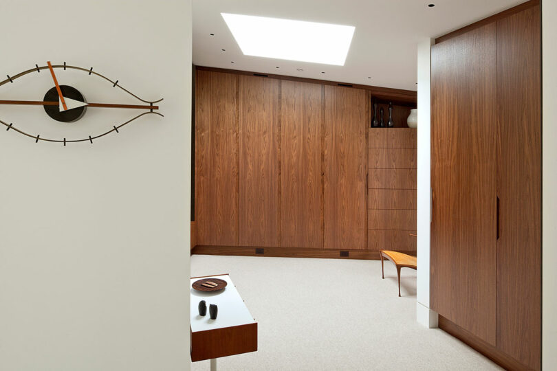 Modern room with wooden cabinets, a wall-mounted clock shaped like an eye, a table, and a skylight. Neutral color palette with brown and white tones.