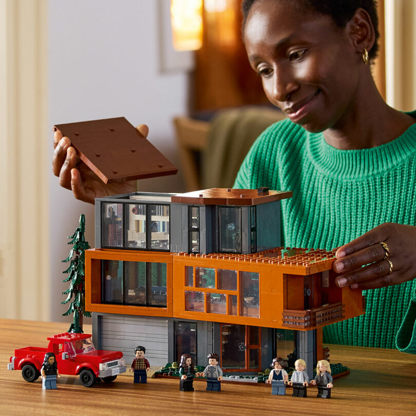 Person in a green sweater assembling a LEGO model of a modern house on a table, with a red toy truck and several LEGO minifigures placed nearby.