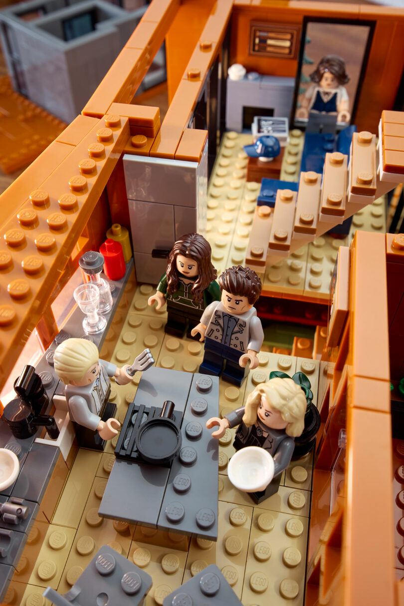 LEGO minifigures in a detailed room setting with a staircase, table, and kitchen area.