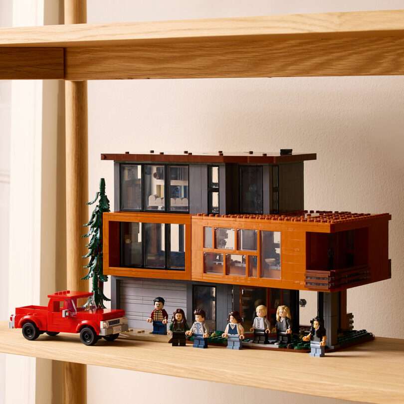 A detailed LEGO model of a modern two-story house with large windows is displayed on a wooden shelf. In front, there is a LEGO red pickup truck and several minifigures.