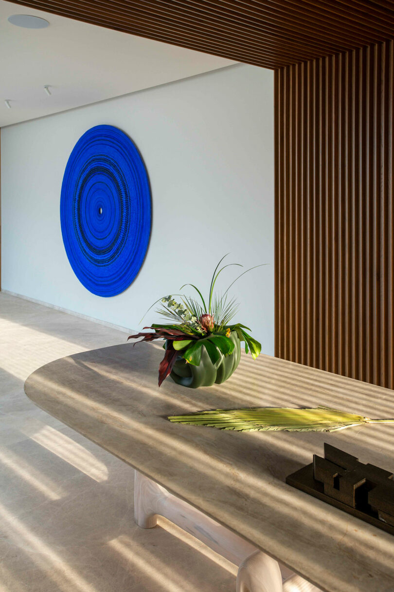 A round blue artwork on a white wall beside vertical wooden panels. In the foreground, a long table with a plant arrangement and decorative items.