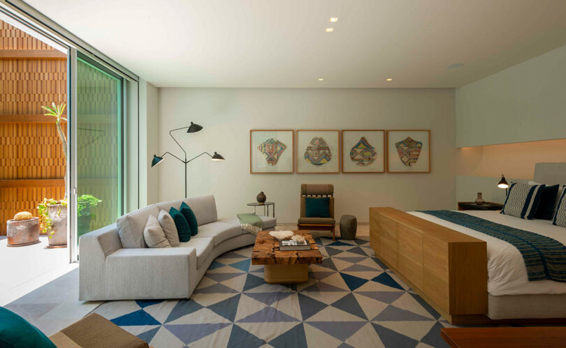 Modern bedroom with a large bed, a gray sectional sofa, and a wooden coffee table on a geometric-patterned rug. Four framed artworks hang on the wall. Sliding glass door leads to a patio.