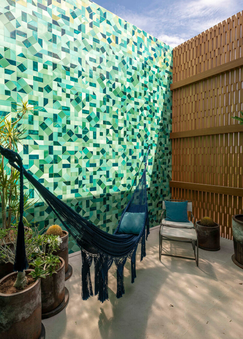 A blue hammock hangs in a corner with a green geometric tile wall and a wooden slat fence. A chair with a cushion and potted plants surround the hammock. The sky is partly cloudy.