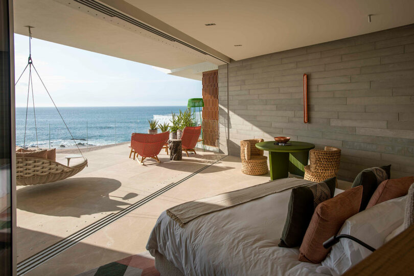 A modern bedroom opens to a spacious balcony with ocean views, featuring a hammock, green table, wicker chairs, and plants.