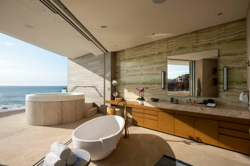 Luxurious bathroom with a freestanding tub and a large vanity, overlooking the ocean through a wide open window. There's a round jacuzzi beside the tub.