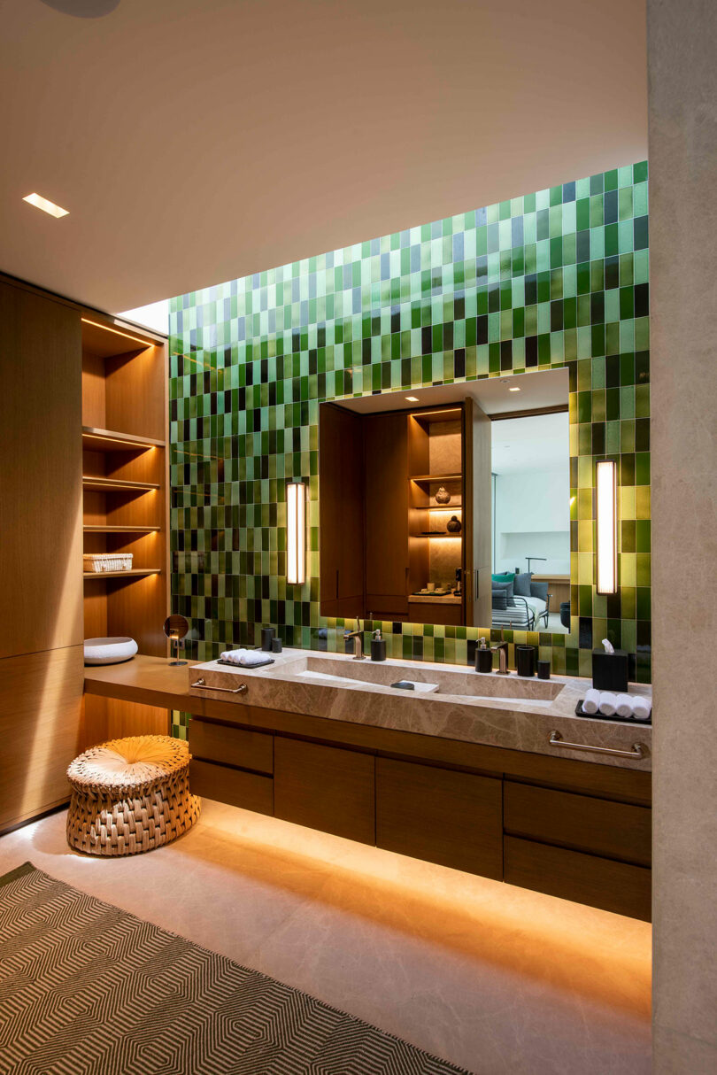 Modern bathroom with green mosaic tile wall, large mirror, and wooden cabinets. Lit wall sconces flank the mirror. A woven basket sits by the marble countertop.