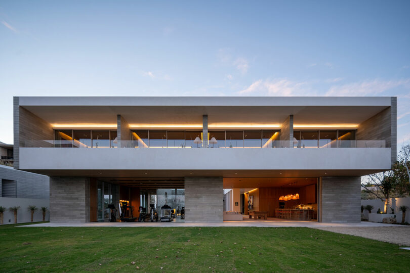 Modern two-story house with large windows, flat roof, and a spacious lawn in front. The evening light highlights the minimalist design.