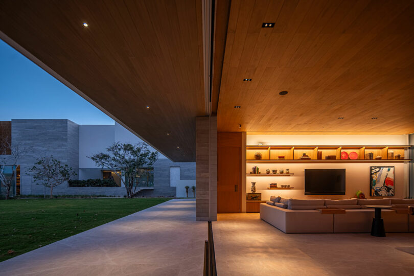 Modern open-concept living room with recessed lighting, a large sectional sofa, and wall-mounted TV. Sliding glass doors open to a spacious patio and lawn area at dusk.