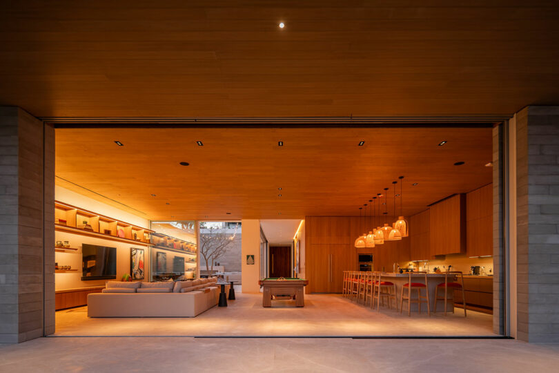 Spacious modern living area with an open layout featuring a kitchen, dining table, seating, and large glass doors leading to an outdoor space, all warmly lit with wooden accents.