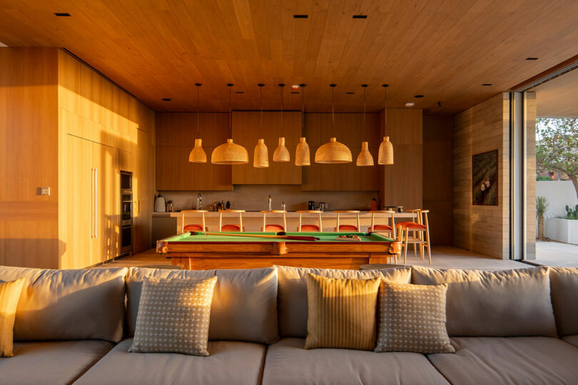 Modern open-plan kitchen and living room with wooden interior, a pool table, pendant lights, and a large beige sofa in the foreground.