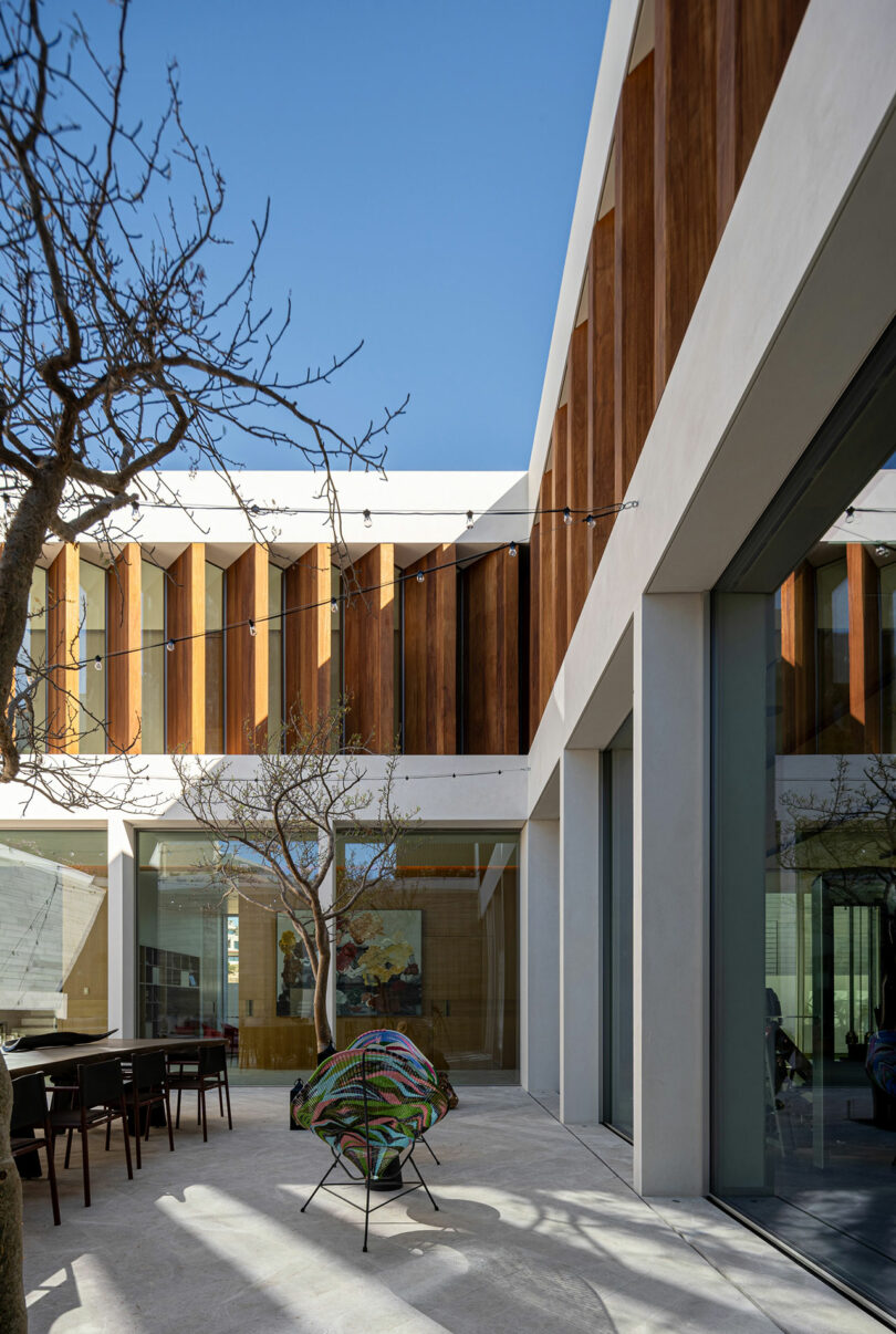 Modern courtyard with a tree and colorful chair, surrounded by a two-story building featuring wood accents and large windows. Blue sky overhead.