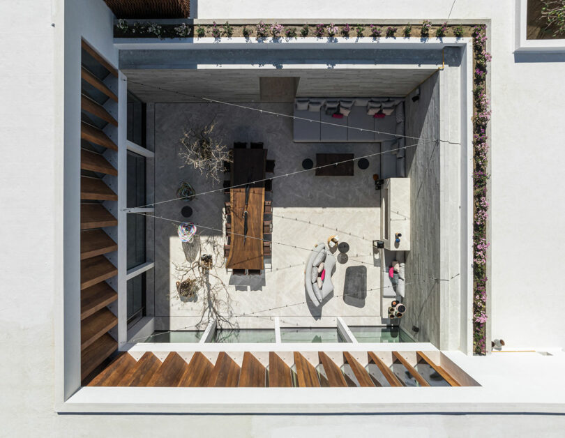 Aerial view of a modern courtyard with a wooden dining table, white sofas, a bare tree, and plant-lined edges.